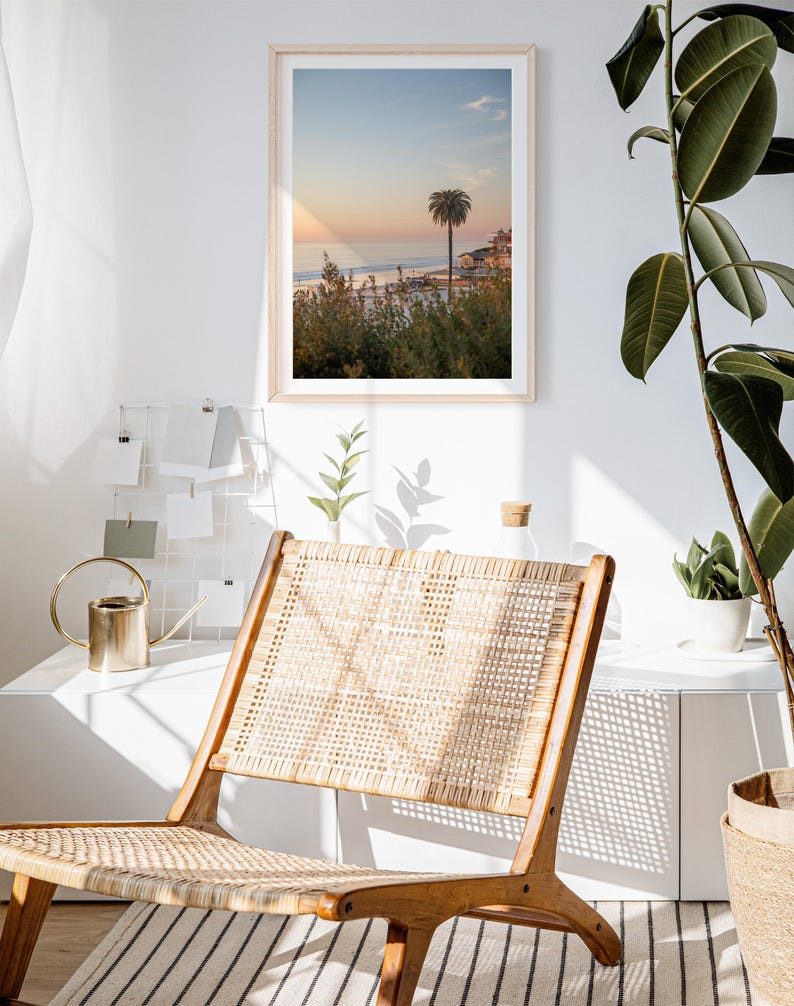 May include: A framed print of a coastal scene with a palm tree at sunset hangs on a white wall. A wooden chair with a woven seat and back sits in front of the wall. A gold watering can and a plant are on a white shelf.