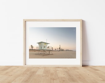 Puede incluir: Fotografía enmarcada de una torre de salvavidas en una playa de arena bajo un cielo despejado. La torre es de color verde claro y blanco con detalles amarillos. La playa está vacía y el cielo es un degradado de azul y naranja.
