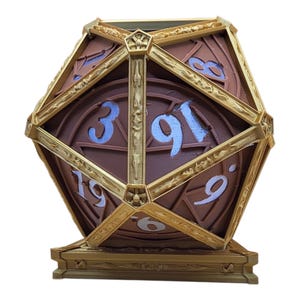 Könnte beinhalten: Ein dekorativer D20-Würfel in Braun und Gold mit blauen Zahlen. Der Würfel ist ein Polyeder mit zwanzig Flächen, die jeweils mit einer Zahl markiert sind. Die goldene Umrandung verleiht dem Würfel ein dekoratives Element.
