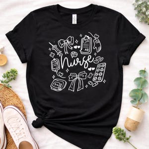 May include: Black t-shirt with a white graphic design featuring the word "Nurse" surrounded by medical-themed illustrations, including a syringe, heart, and pills. The shirt has short sleeves and a crew neck.