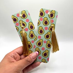 May include: Two avocado-themed bookmarks with gold tassels. The bookmarks feature a repeating pattern of smiling avocados and pink flowers on a light green background.