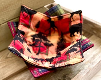 May include: A set of three red, black, and green plaid fabric bowl cozies with a woodland animal print. The cozies are folded and stacked in a wooden tray.