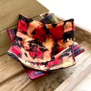 Puede incluir: Un juego de tres posavasos de tela a cuadros rojos, negros y verdes con estampado de animales del bosque. Los posavasos están doblados y apilados en una bandeja de madera.