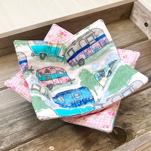 Puede incluir: Un juego de dos calientaplatos de tela acolchados con un tema de caravanas vintage. Los calientaplatos están hechos de tela blanca con un estampado floral verde, azul y rosa y presentan una variedad de ilustraciones de caravanas coloridas. Los calientaplatos están forrados con tela de cuadros rosa y blanca.