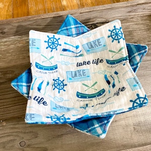 Puede incluir: Dos agarraderas de olla de tela azul y blanca con un tema de lago. Las agarraderas tienen un fondo blanco con ilustraciones azules y verdes de barcos, remos y un volante. El texto "lake life" está impreso en azul en las agarraderas.