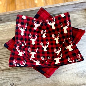 Puede incluir: Calientaplatos de búfalo rojo y negro con siluetas de ciervos blancas. El calientaplatos está doblado en forma de estrella y está sobre una superficie de madera.