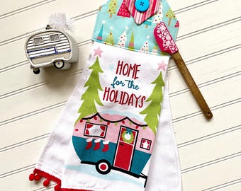 Puede incluir: Toalla de cocina blanca con una caravana roja, blanca y azul con el texto "Home for the Holidays". La toalla tiene pompones rojos alrededor del borde inferior y un lazo de colgar a rayas rosa y blanco con un botón azul.