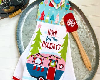 Puede incluir: Un paño de cocina blanco para colgar con borde de pompones rojos, que presenta un gráfico de una caravana rosa y azul con el texto "HOME for the HOLIDAYS". La parte superior es de tela azul claro con árboles coloridos y un botón azul.
