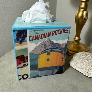 May include: A blue and white tissue box cover with a vintage travel poster design featuring a train, mountains, and the text "Experience the CANADIAN ROCKIES".