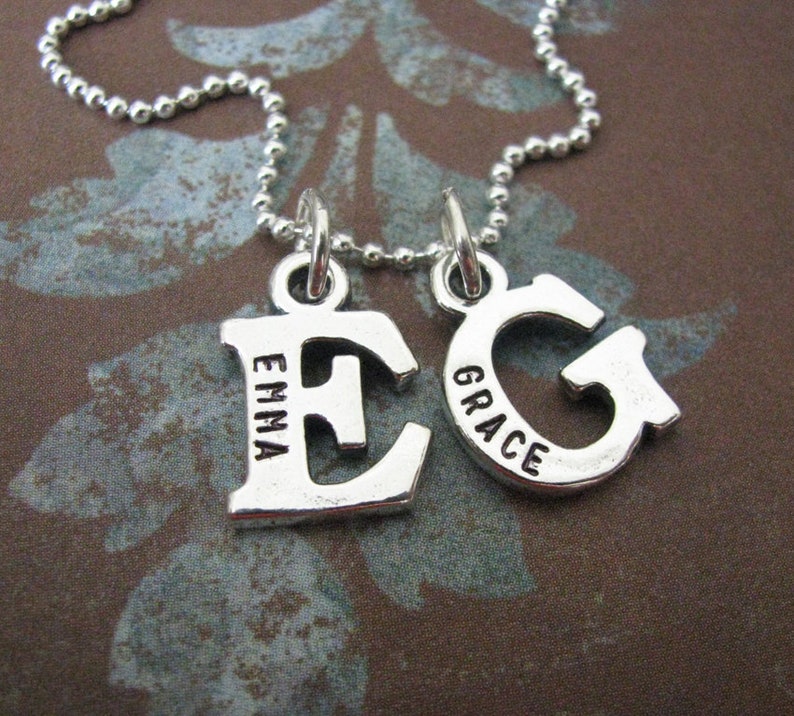 Peut inclure: Un collier en argent avec deux pendentifs, l'un avec la lettre "E" et le mot "Emma" grav&eacute; dessus, et l'autre avec la lettre "G" et le mot "Grace" grav&eacute; dessus.