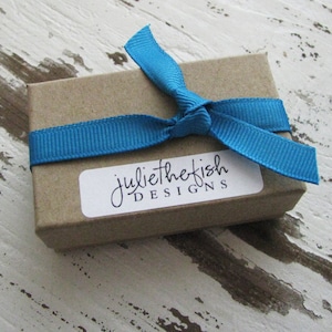 Peut inclure: Une petite bo&icirc;te brune avec un ruban bleu nou&eacute; autour. La bo&icirc;te a une &eacute;tiquette blanche avec le texte "juliethefish DESIGNS" dessus.