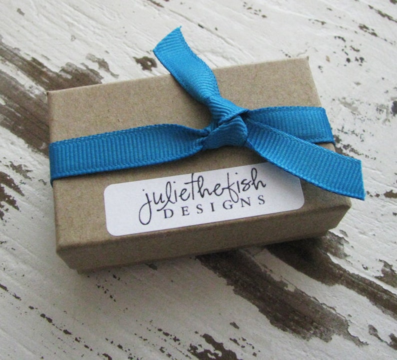 Peut inclure: Une petite bo&icirc;te brune avec un ruban bleu nou&eacute; autour. La bo&icirc;te a une &eacute;tiquette blanche avec le texte "juliethefish DESIGNS" dessus.