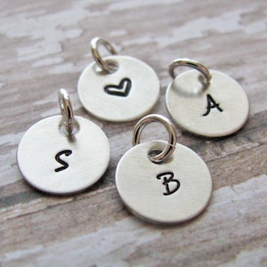 Peut inclure: Quatre petits charms ronds argentés avec une boucle en haut. L'un des charms a un cœur, et les autres ont les lettres "S", "A" et "B" en noir. Ces charms sont parfaits pour la création de bijoux.