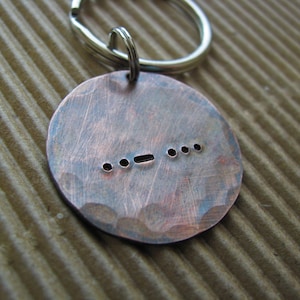 May include: A copper keychain with a hammered finish and a series of dots and dashes engraved on the front. The keychain is attached to a silver key ring.