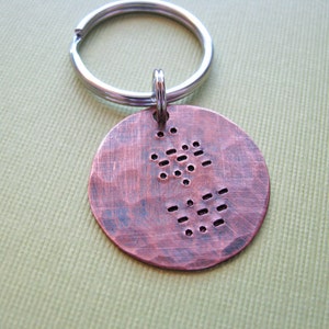 May include: A round, copper-colored keychain tag with a hammered texture. The tag features a series of punched holes arranged in a pattern, and is attached to a silver-colored key ring.