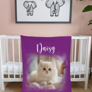 K&ouml;nnte beinhalten: Eine violette Babydecke mit dem Namen "Daisy" in wei&szlig;er Schrift. Die Decke zeigt eine flauschige wei&szlig;e Katze in einem weichen, verschwommenen Hintergrund. &Uuml;ber dem Kinderbett zeigt ein gerahmtes Bild zwei Elefantenzeichnungen und ein rotes Herz.