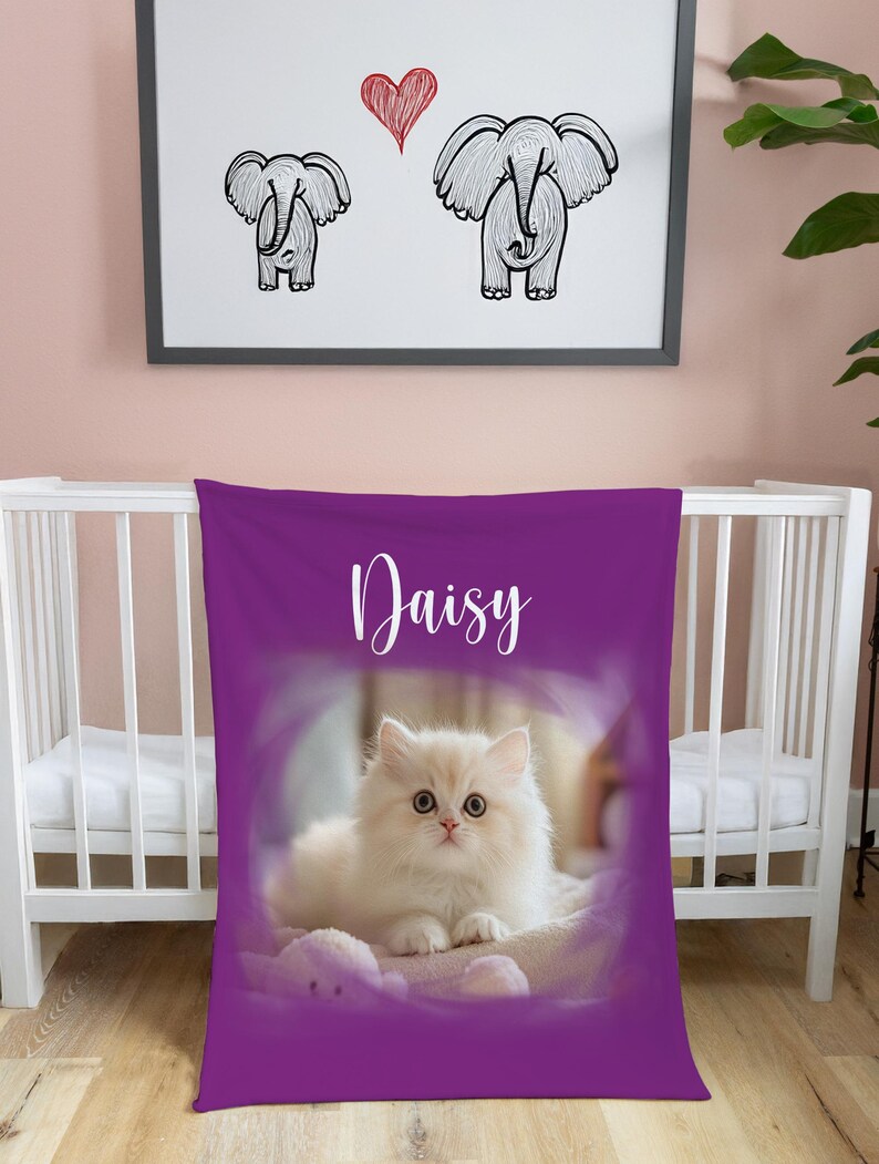 K&ouml;nnte beinhalten: Eine violette Babydecke mit dem Namen "Daisy" in wei&szlig;er Schrift. Die Decke zeigt eine flauschige wei&szlig;e Katze in einem weichen, verschwommenen Hintergrund. &Uuml;ber dem Kinderbett zeigt ein gerahmtes Bild zwei Elefantenzeichnungen und ein rotes Herz.