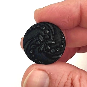 May include: A black, round button with a floral design. The button has a raised, textured pattern with a central flower and swirling accents. Small, dark embellishments are scattered around the design. The button is held between fingers.