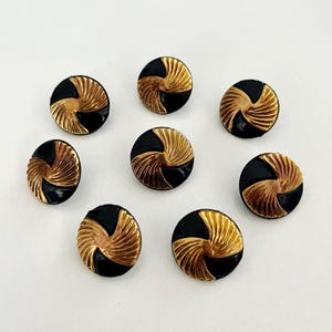 May include: Eight round buttons with a black and gold design. The buttons have a black base with a gold, fan-like pattern. The buttons are arranged in a circular pattern on a white surface. These buttons are ideal for sewing and craft projects.