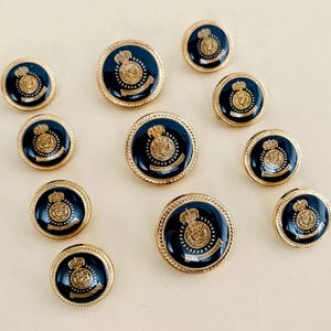 Puede incluir: Un conjunto de botones azul marino y dorado con un diseño de escudo real. Los botones tienen un borde dorado en forma de cuerda y un acabado brillante. Se muestran varios tamaños sobre una superficie color crema.