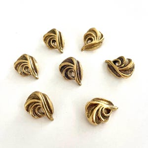 Può includere: Sette bottoni antichi color oro con un design a spirale stilizzato. Ogni bottone presenta una spirale centrale circondata da elementi curvi e strutturati, creando un effetto tridimensionale. I bottoni sono disposti su una superficie bianca.