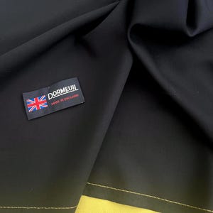 Peut inclure: Tissu noir avec une couture en fil doré. Une étiquette avec le texte "Dormeuil Made in England" est attachée au tissu.