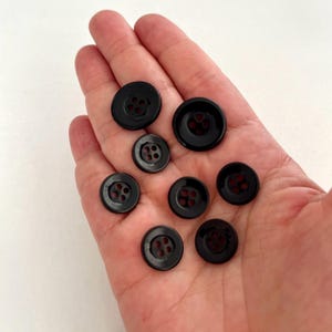 May include: A collection of seven round, black buttons of varying sizes, each featuring a smooth, glossy finish and four small holes in the center for attachment. These circular fasteners are arranged in a cluster, displaying a consistent dark color.