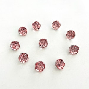 Pode incluir: Uma coleção de dez contas de vidro rosa em forma de rosa. Cada conta tem um design espiralado detalhado e estão espalhadas sobre uma superfície branca. As contas têm aproximadamente 1,3 cm de diâmetro, ideais para fazer joias ou projetos de artesanato.