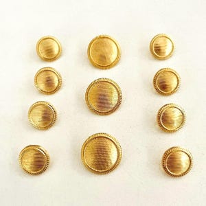 May include: A collection of gold-colored buttons with a textured, ridged surface and a rope-like border. The buttons vary in size and are arranged on a white surface. These decorative buttons are suitable for sewing and craft projects.