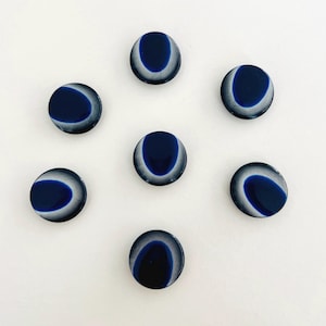 Puede incluir: Siete botones redondos de color azul oscuro con un diseño de media luna blanco y azul claro. Los botones están dispuestos en un patrón circular sobre un fondo blanco. Los botones son probablemente para coser o manualidades.