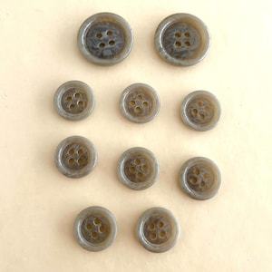 May include: A collection of eleven gray buttons with four holes each. The buttons vary in size, with a marbled appearance. The buttons are arranged on a light beige surface.