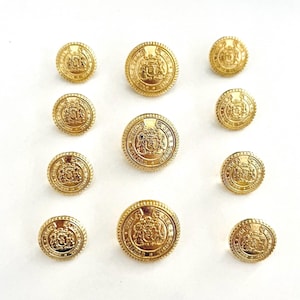 May include: A collection of ten round gold-toned metal buttons arranged in three rows. Each button features a raised embossed crest design in the centre, surrounded by a circular border with text and a textured, rope-like edge detail.