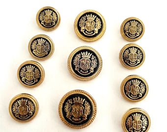 May include: A collection of ten round buttons in varying sizes, each featuring a black center with a gold-toned embossed crest design. The crest depicts a crown above a shield flanked by laurel leaves, all surrounded by a gold-toned rope-textured rim.