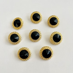 May include: Eight round buttons with a gold-colored, textured border and a black, faceted center. The buttons are arranged on a white surface, showcasing their decorative design. These buttons are suitable for sewing and craft projects.