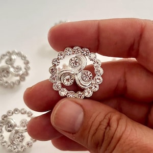 May include: A circular silver-toned button featuring a central triskelion design embellished with clear rhinestones. The outer edge of the button is surrounded by a ring of small, clear, round rhinestones, creating a decorative metallic accessory.