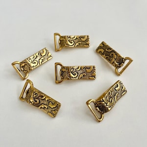 Puede incluir: Seis cierres antiguos de color latón con adornos de volutas. Cada cierre presenta un cuerpo rectangular con un patrón decorativo y una anilla en D en un extremo. Los cierres están dispuestos en un patrón circular sobre un fondo blanco.