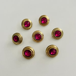 Może przedstawiać: Siedem złotych guzików z wypukłym, okrągłym wzorem. Każdy guzik ma w środku żywy, okrągły, magentowy klejnot. Guziki ułożone są na białej powierzchni.