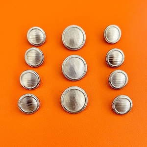 May include: A collection of ten round silver-toned buttons arranged in three columns. Each button features a textured, horizontally lined center surface surrounded by a thin, raised, rope-like border detail along the outer edge.