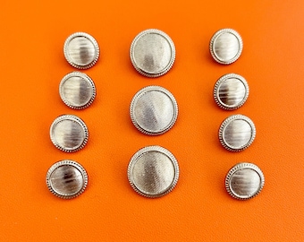 May include: A collection of ten round silver-toned buttons arranged in three columns. Each button features a textured, horizontally lined center surface surrounded by a thin, raised, rope-like border detail along the outer edge.