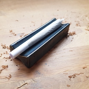 Peut inclure: Un plateau de roulage en céramique bleu foncé avec un joint pré-roulé blanc à l'intérieur. Le plateau est sur une surface en bois avec des copeaux de tabac brun éparpillés autour.