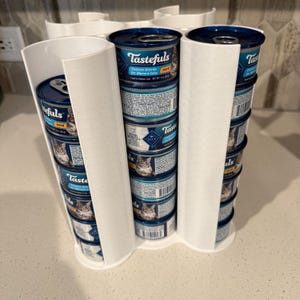 May include: A white plastic vertical cat food can dispenser holding multiple stacked 3-ounce cans of Tastefuls Chicken Entrée for Mature Cats. The dispenser features curved channels that organize and display the blue and white metal cans.
