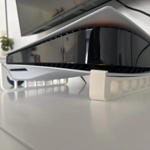 Könnte beinhalten: Eine weiße und schwarze Spielekonsole mit passendem weißem Ständer. Die Konsole hat ein schlankes, geschwungenes Design mit einem glänzenden schwarzen Mittelteil. Der Ständer ist rechteckig mit einer Reihe offener Schlitze zur Belüftung.