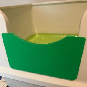May include: A close-up of a white and green storage bin. The bin has a bright green front panel with a curved top. The interior of the bin is a light green color. The bin is likely used for organizing items.