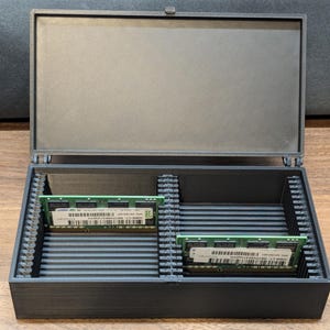 May include: A black, hinged storage box with two green RAM sticks inside. The box is open, revealing the interior with slots for the RAM. The lid is closed and has a textured surface. The box is on a wooden surface.