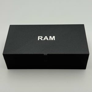 May include: A black rectangular box with the word "RAM" in white capital letters centered on the top. The box has a textured surface and a small indentation on the front.