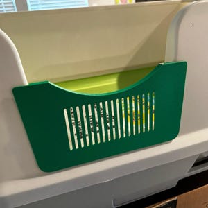 May include: A white and green cat litter tray with a green scoop and a green grate. The litter tray has a removable top and a built-in scoop holder.