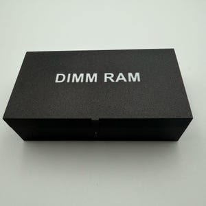 May include: A black rectangular box with the white text "DIMM RAM" on the top. The box has a textured surface and a hinged lid. The background is a light, neutral color.