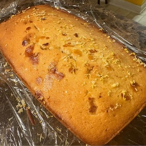 May include: A golden-brown loaf of bread, possibly a cake or quick bread, sits on a surface covered in clear plastic wrap. The bread has a slightly textured surface and is sprinkled with yellow crumbs. The overall appearance suggests a homemade baked good.