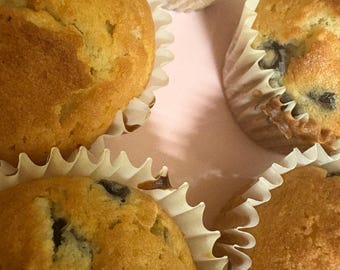 Può includere: Primo piano di cinque muffin dorati in pirottini di carta bianchi, disposti su una superficie rosa chiaro. Alcuni muffin hanno bacche blu scuro o nere visibili. I muffin sembrano appena sfornati.