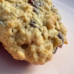 Puede incluir: Primer plano de una galleta de avena y pasas de color marrón dorado sobre un plato rosa. La galleta tiene una superficie texturizada con avena visible y pasas oscuras. Los bordes son ligeramente crujientes.